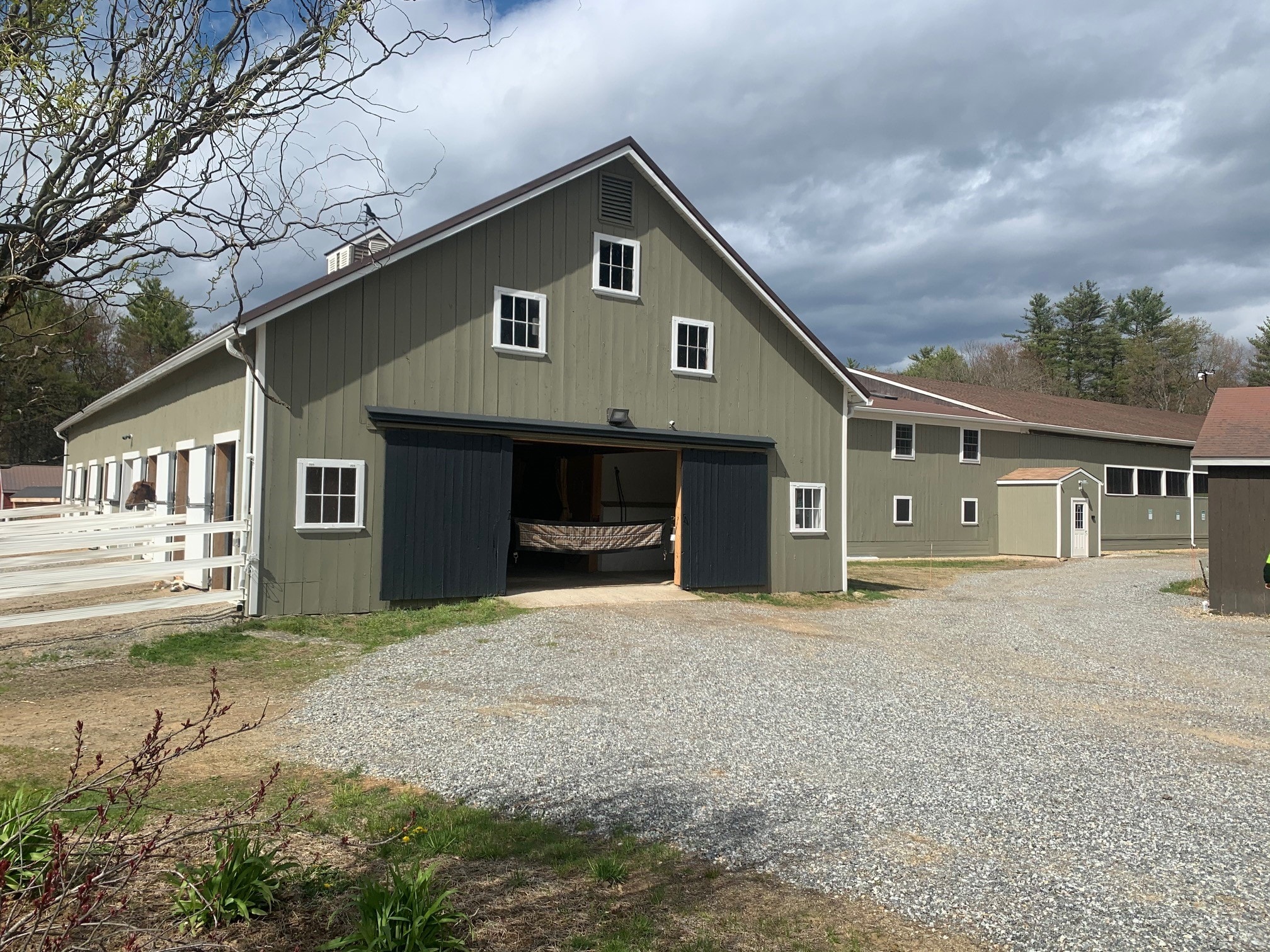 Century Farm Renovations completed exterior with new sage-green board-and-batten siding, white trim, dark sliding barn doors, gravel drive, and white paddock fence — Belmont, NH