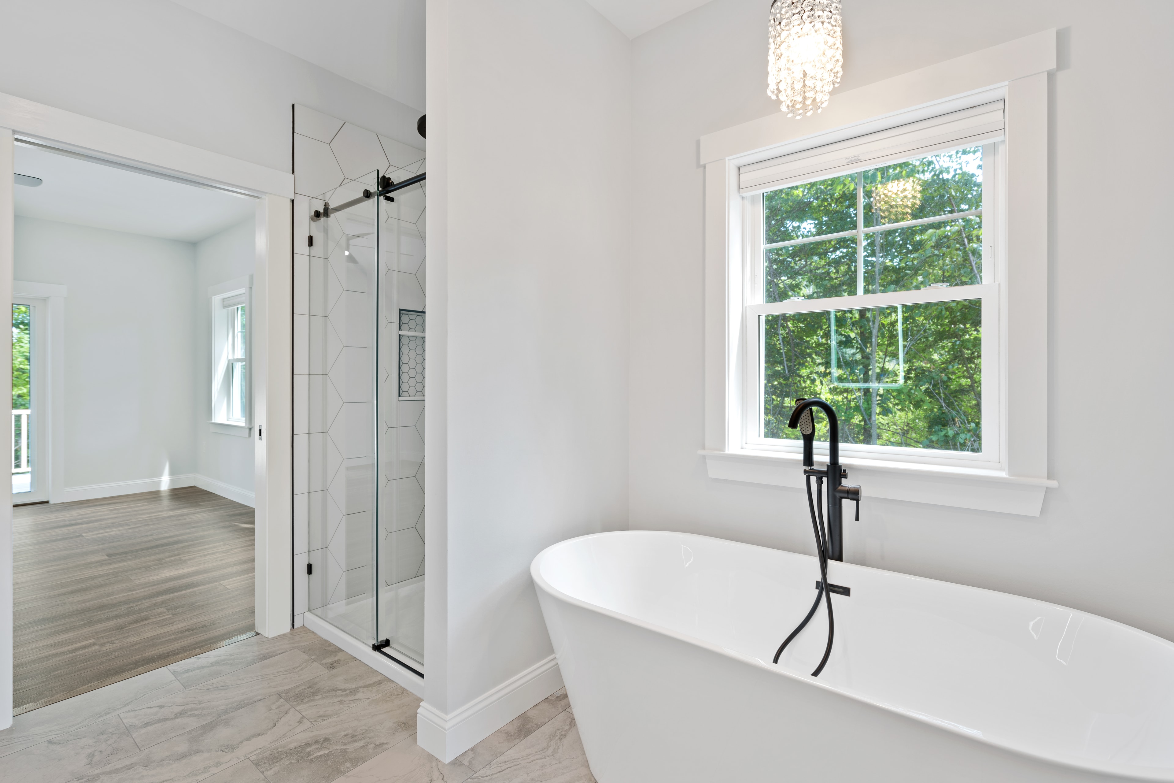 Clover Ridge Home master bath with freestanding soaking tub, hexagon tile walk-in shower, black fixtures, and crystal chandelier — Meredith, NH