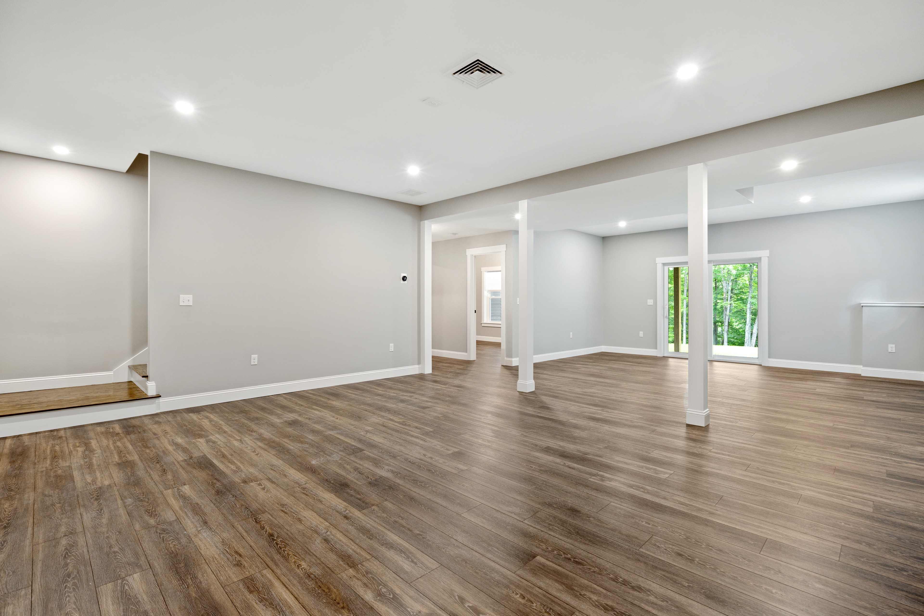 Clover Ridge Home finished lower level with open floor plan, support columns, wide-plank floors, and walkout to wooded yard — Meredith, NH