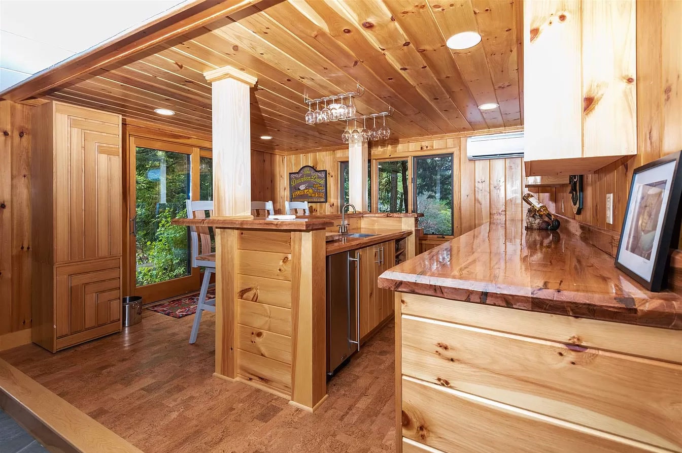 Dew Duck House knotty pine kitchen with live-edge wood countertops, wine glass rack, stainless undermount sink, mini fridge, and forest-view windows — Meredith, NH
