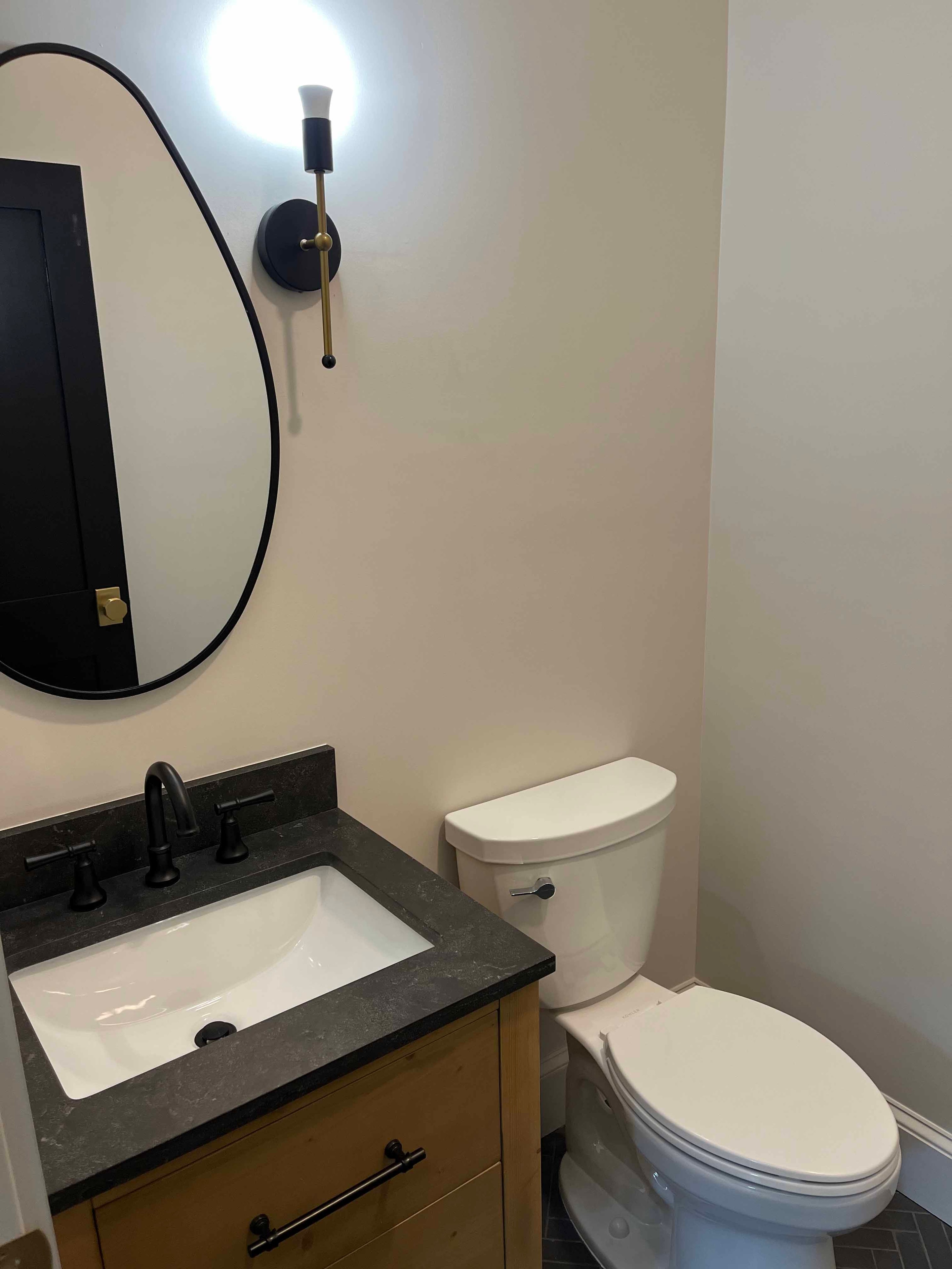 Holderness Home powder bath with wood vanity, dark granite countertop, oval black mirror, and brass sconce
