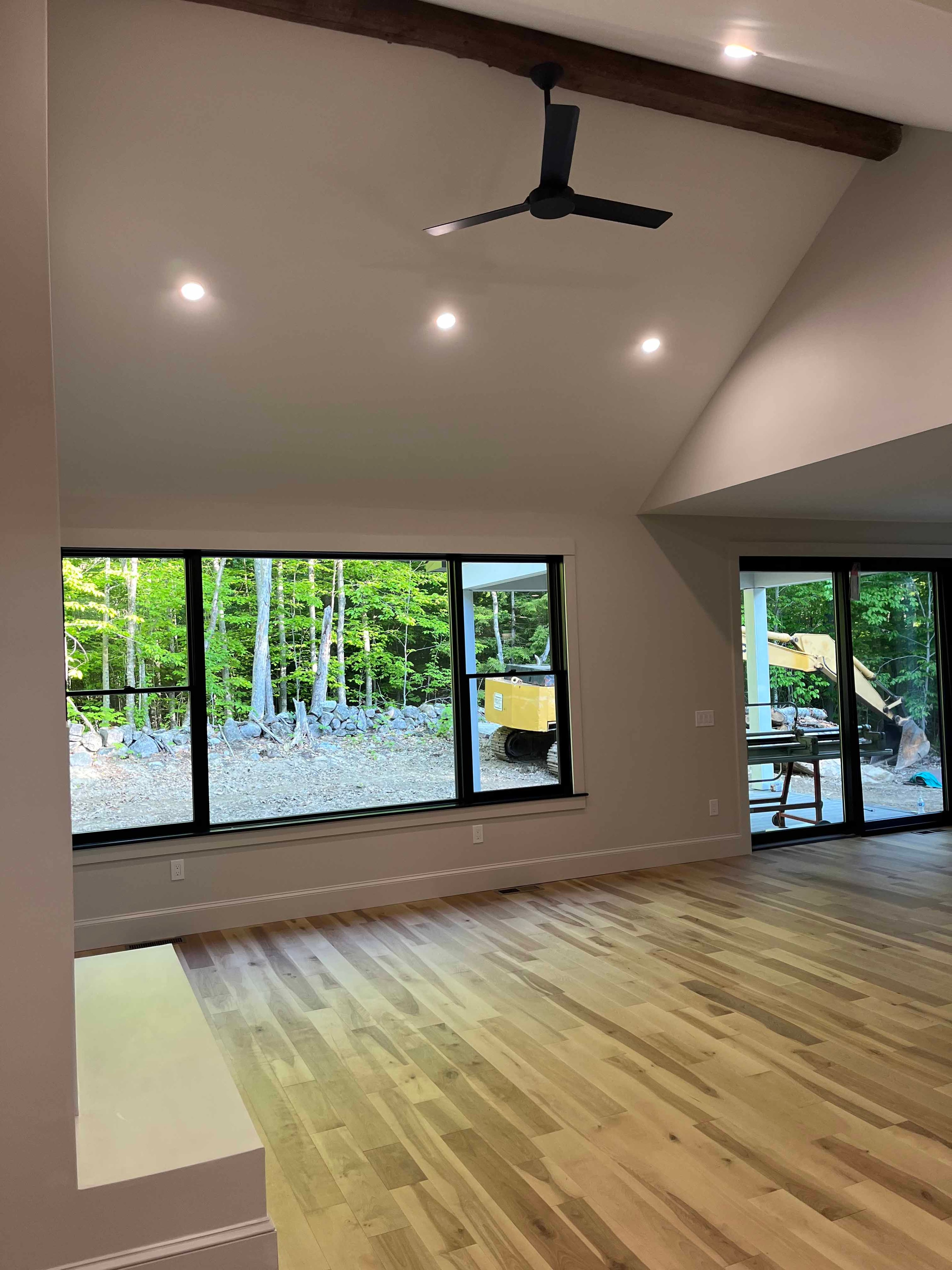 Holderness Home great room with vaulted ceiling, exposed wood beam, black ceiling fan, and black-framed windows overlooking woods