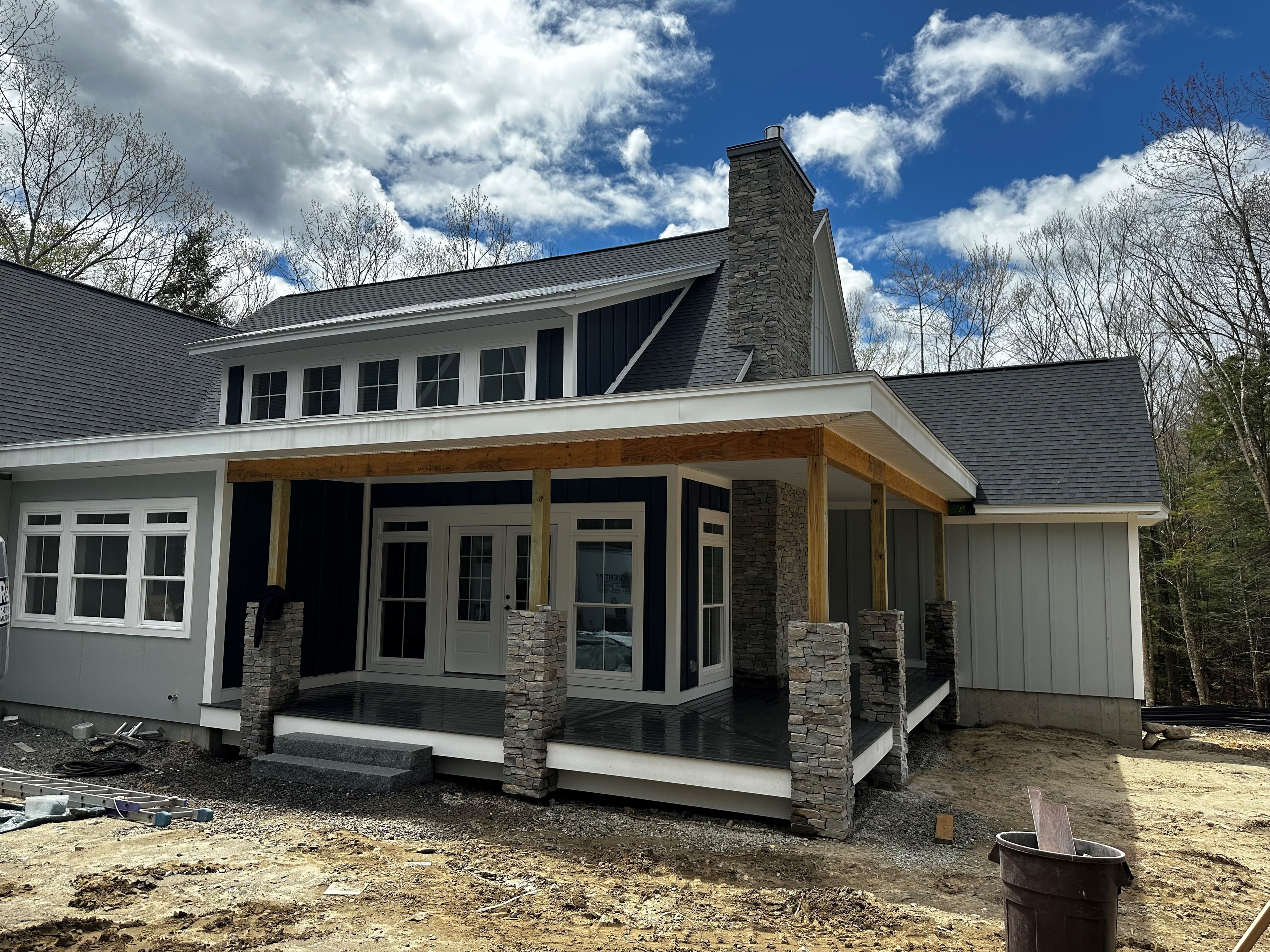 Home in Progress exterior with navy board-and-batten siding, stone porch columns, timber beams, and stone chimney — Moultonborough, NH
