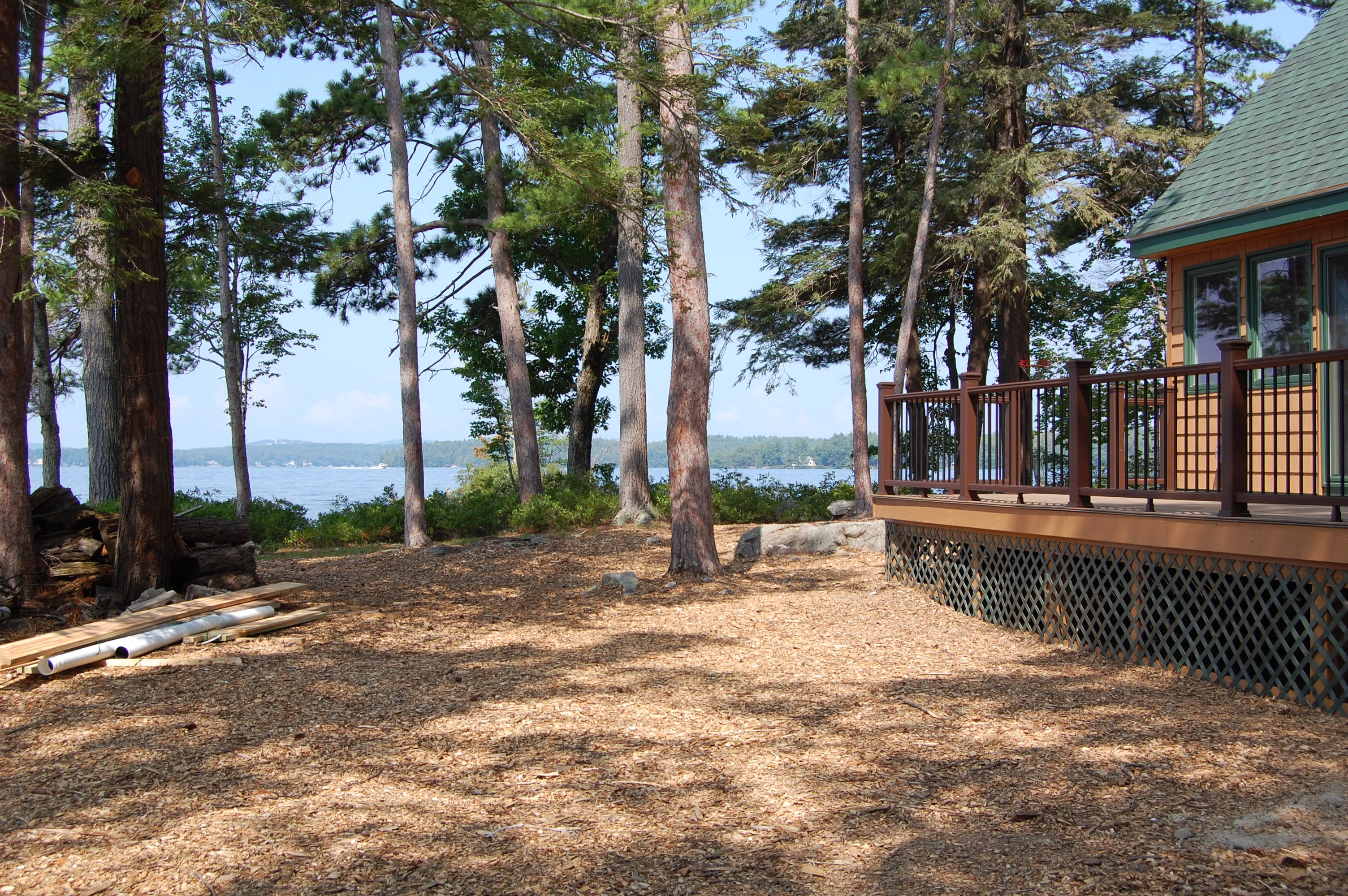 Island Retreat completed lakeside grounds with new cedar deck, dark railing, tall pines, and open lake views — Welch Island, Laconia, NH