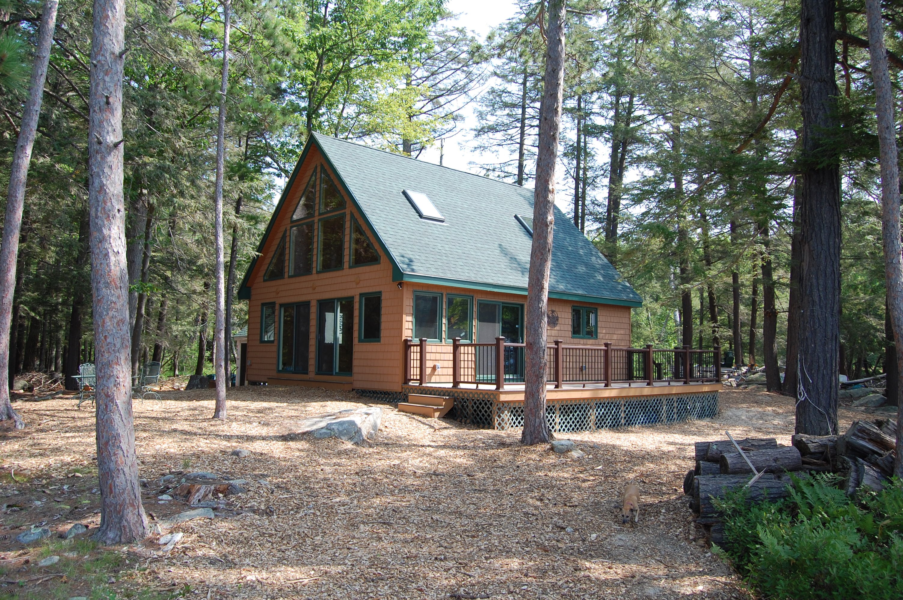 Island Retreat completed cedar shake A-frame exterior with green roof, large gable windows, wraparound deck, and dark railing surrounded by tall pines — Welch Island, Laconia, NH