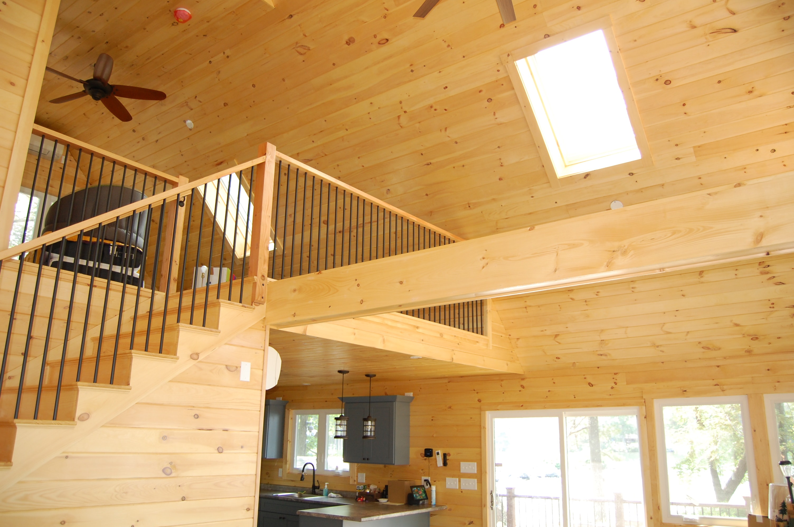 Island Retreat open great room with knotty pine walls and vaulted ceiling, loft with black metal railing, staircase, and sliding glass doors to deck — Welch Island, Laconia, NH