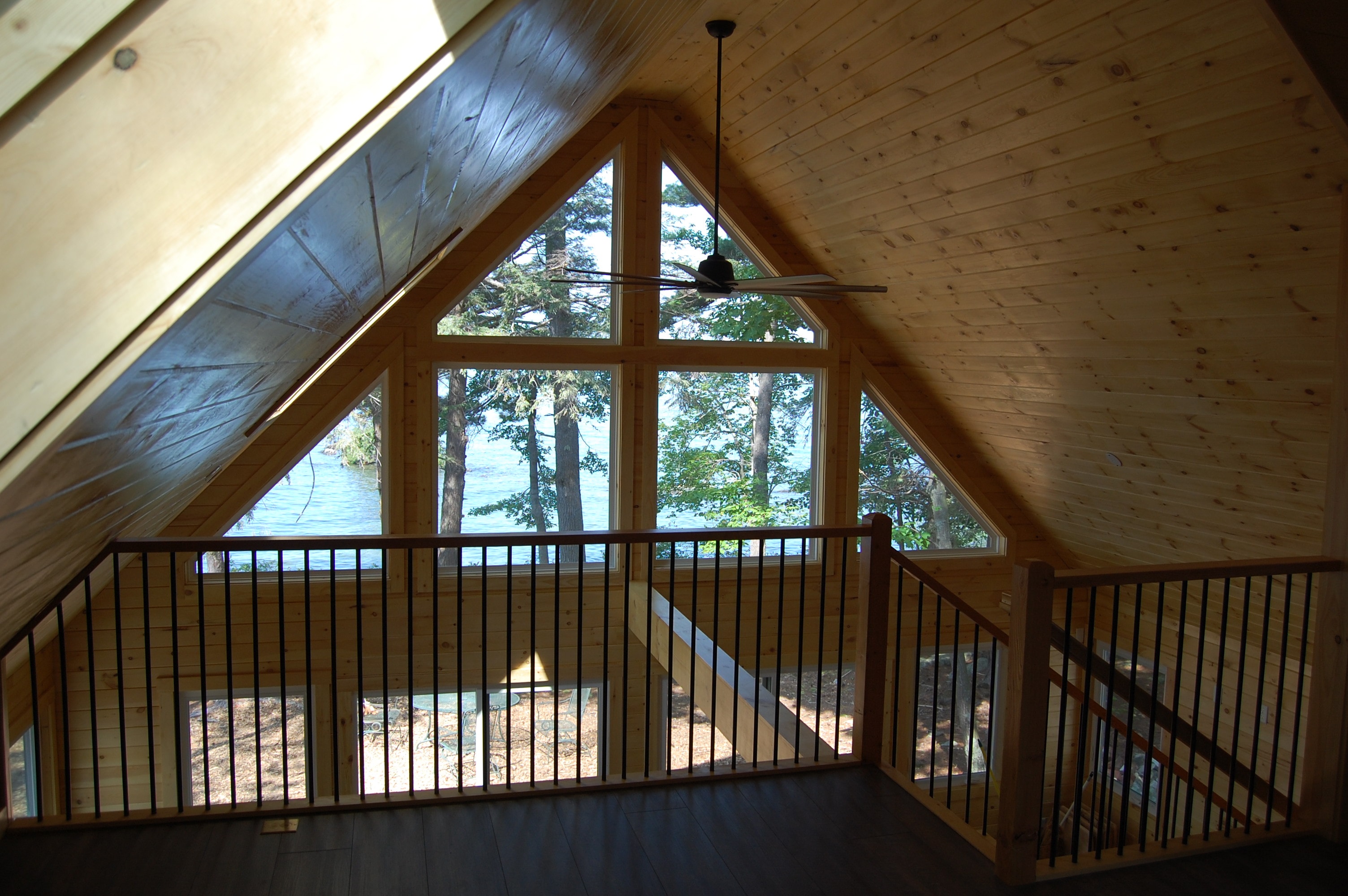 Island Retreat loft with knotty pine vaulted ceiling, skylight, black metal railing, ceiling fan, and lake views through tall A-frame gable windows — Welch Island, Laconia, NH