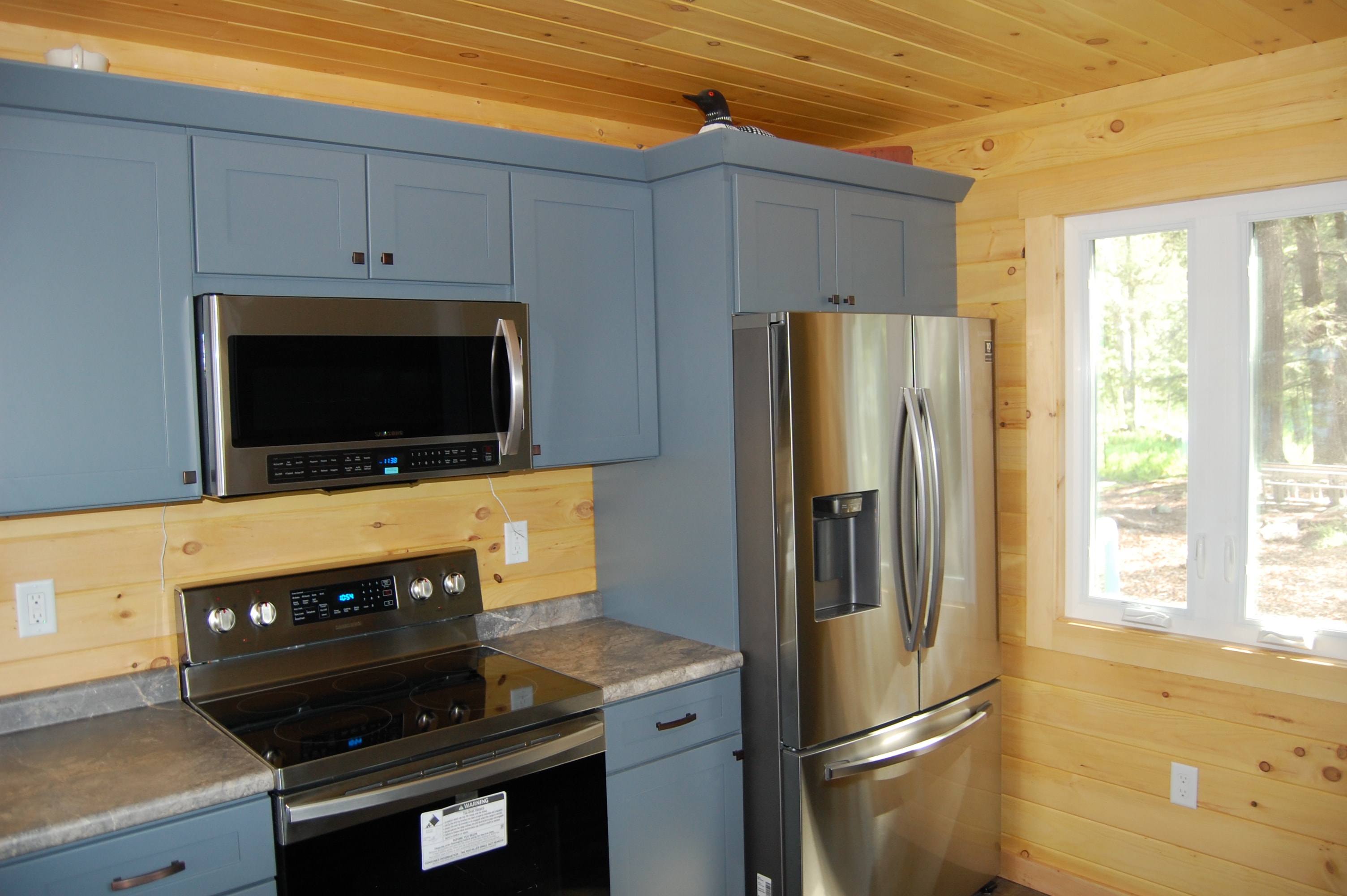 Island Retreat gray-blue kitchen with stainless range, over-range microwave, French door refrigerator, and knotty pine walls and ceiling — Welch Island, Laconia, NH