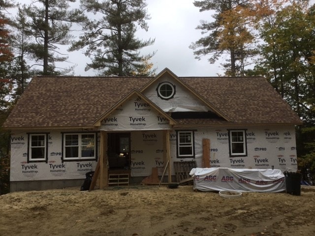 Lake Kanasatka Home front exterior framing with Tyvek wrap, round dormer window, roofing complete, and autumn trees — Moultonborough, NH