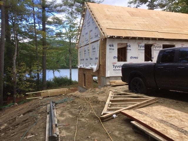 Lake Kanasatka Home side exterior framing with Tyvek wrap, lake view through trees, and construction truck on site — Moultonborough, NH