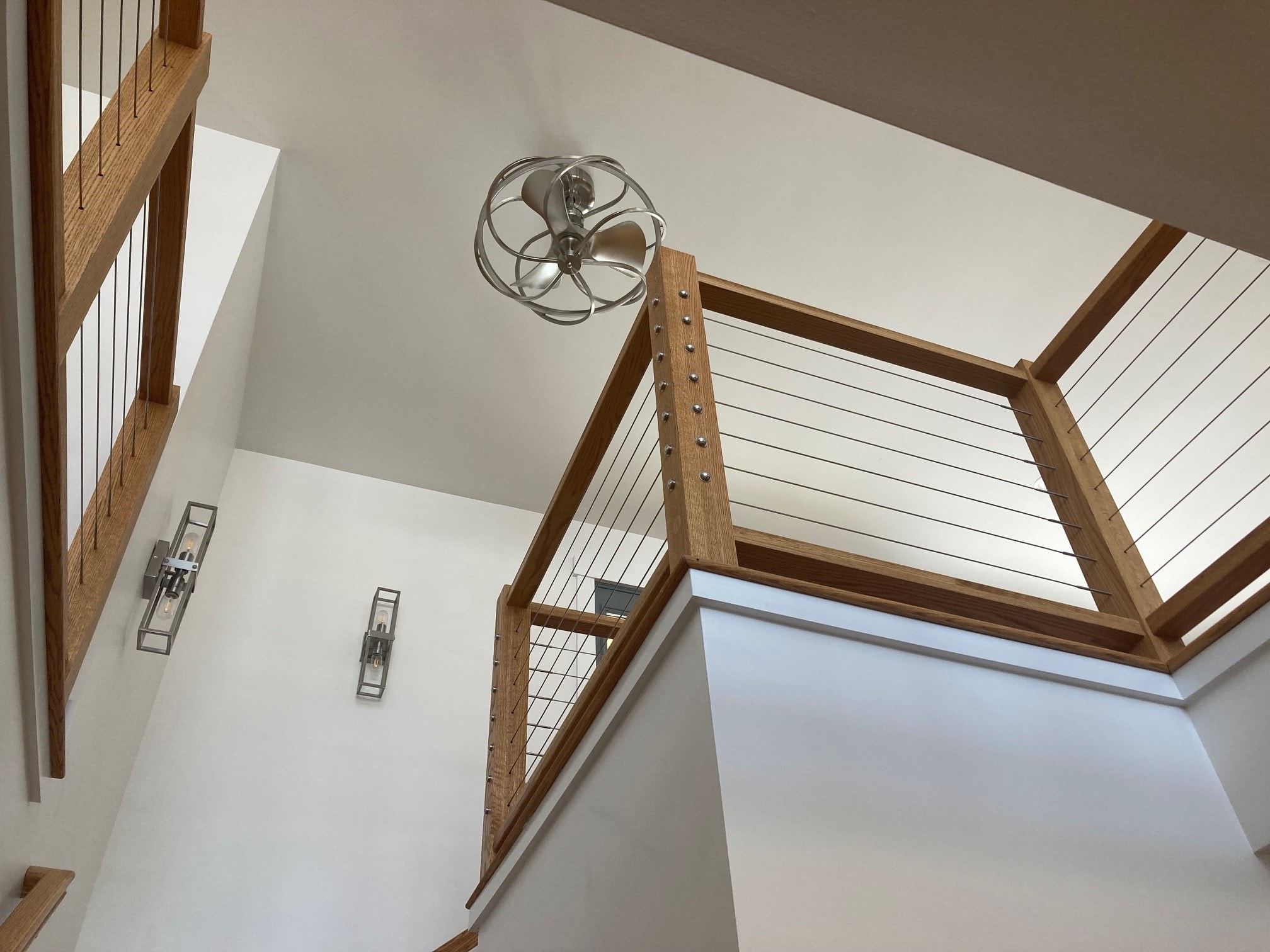 Lake Kanasatka Home stairwell looking up at oak cable-rail landing, orb ceiling fan, and wall sconces — Moultonborough, NH