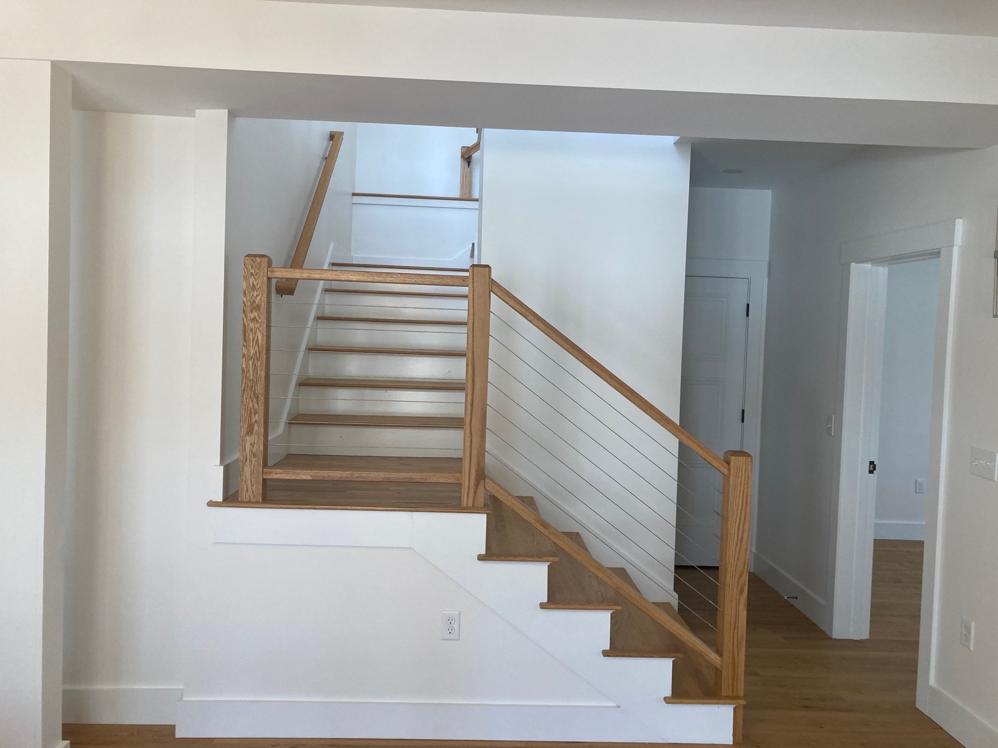 Lake Kanasatka Home oak staircase with cable-rail balusters, white risers, and view to first-floor hallway — Moultonborough, NH