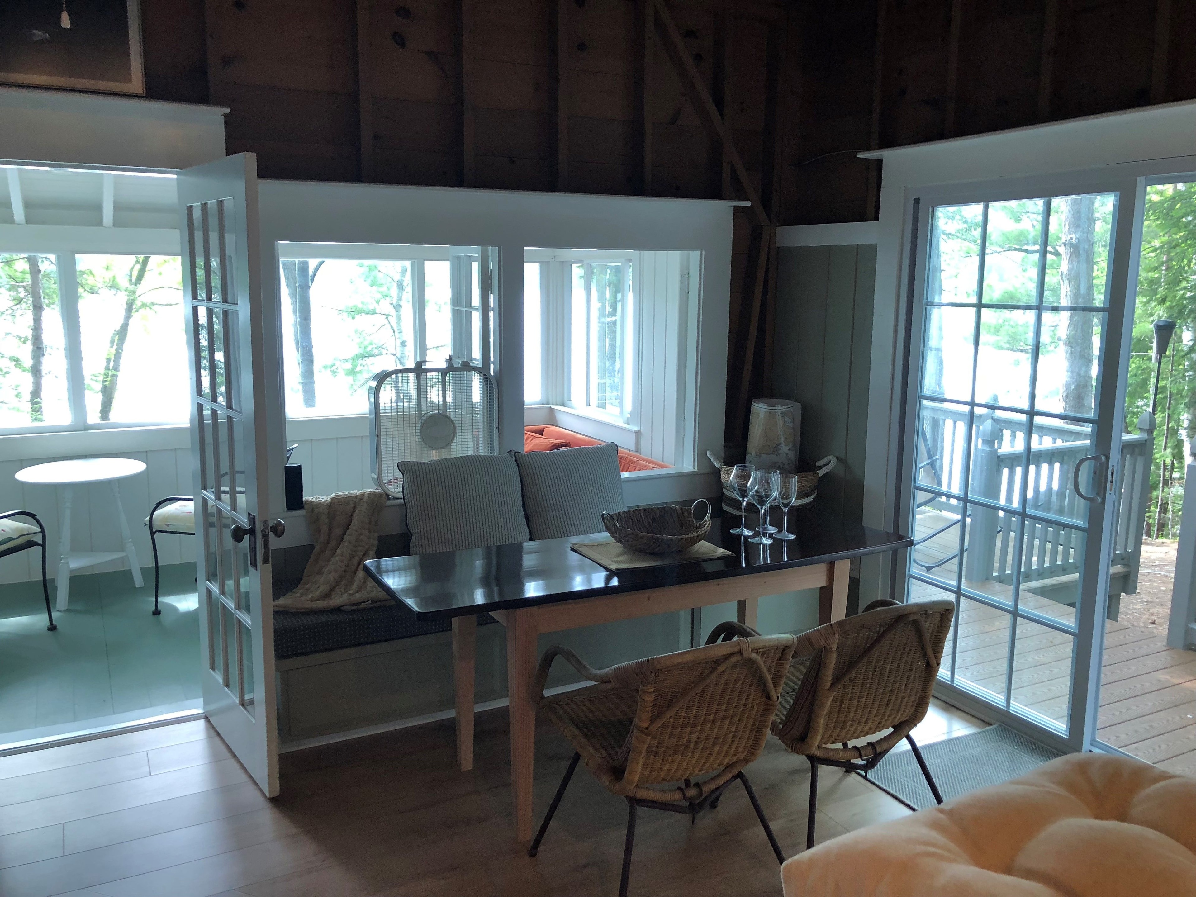 Loon Song Cottage dining area with dark table, wicker chairs, window seat, and French doors opening to wooded deck — Meredith, NH