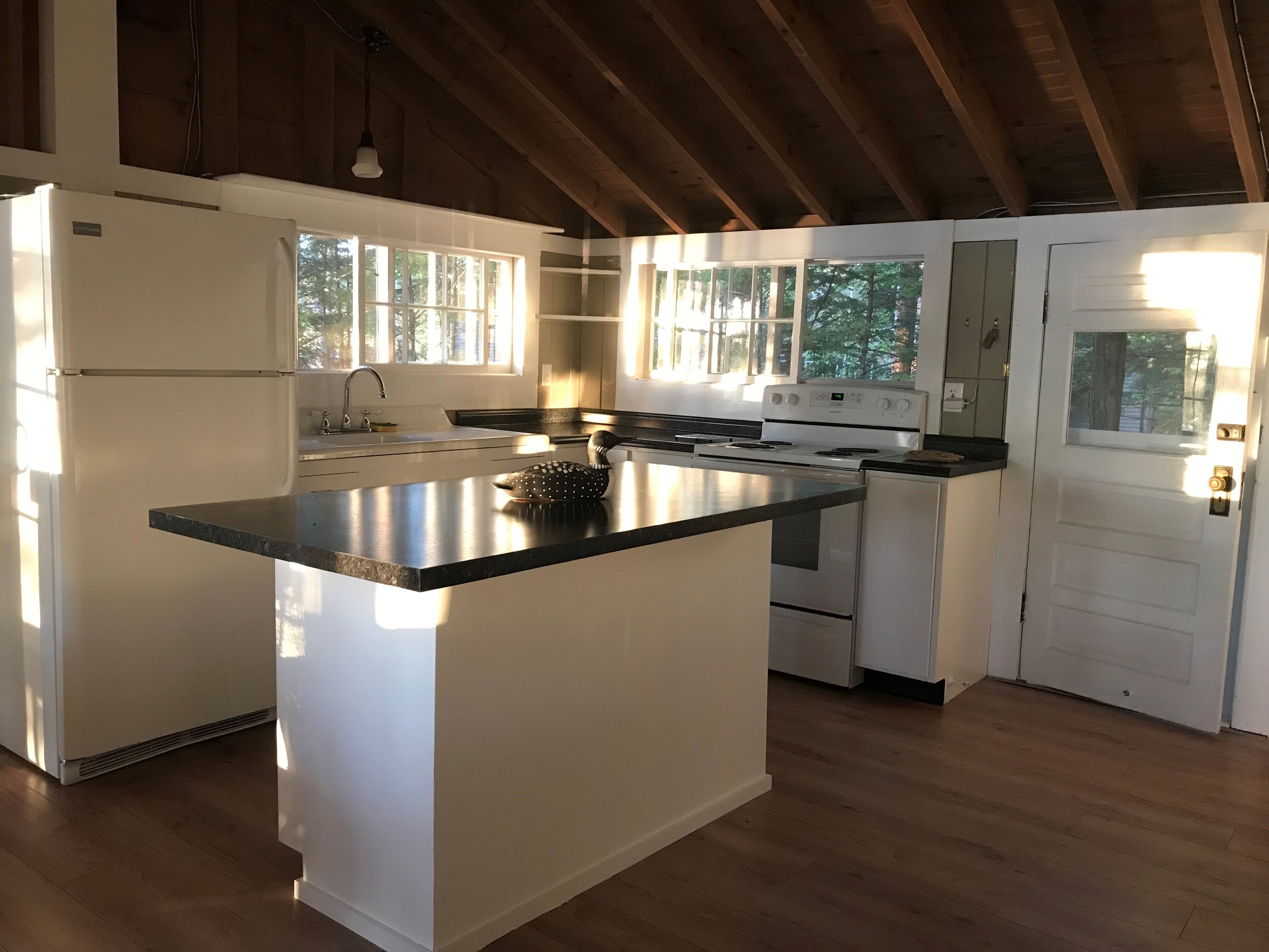 Loon Song Cottage kitchen with white cabinets, black granite island and countertops, white appliances, exposed beam ceiling, and forest-view windows — Meredith, NH