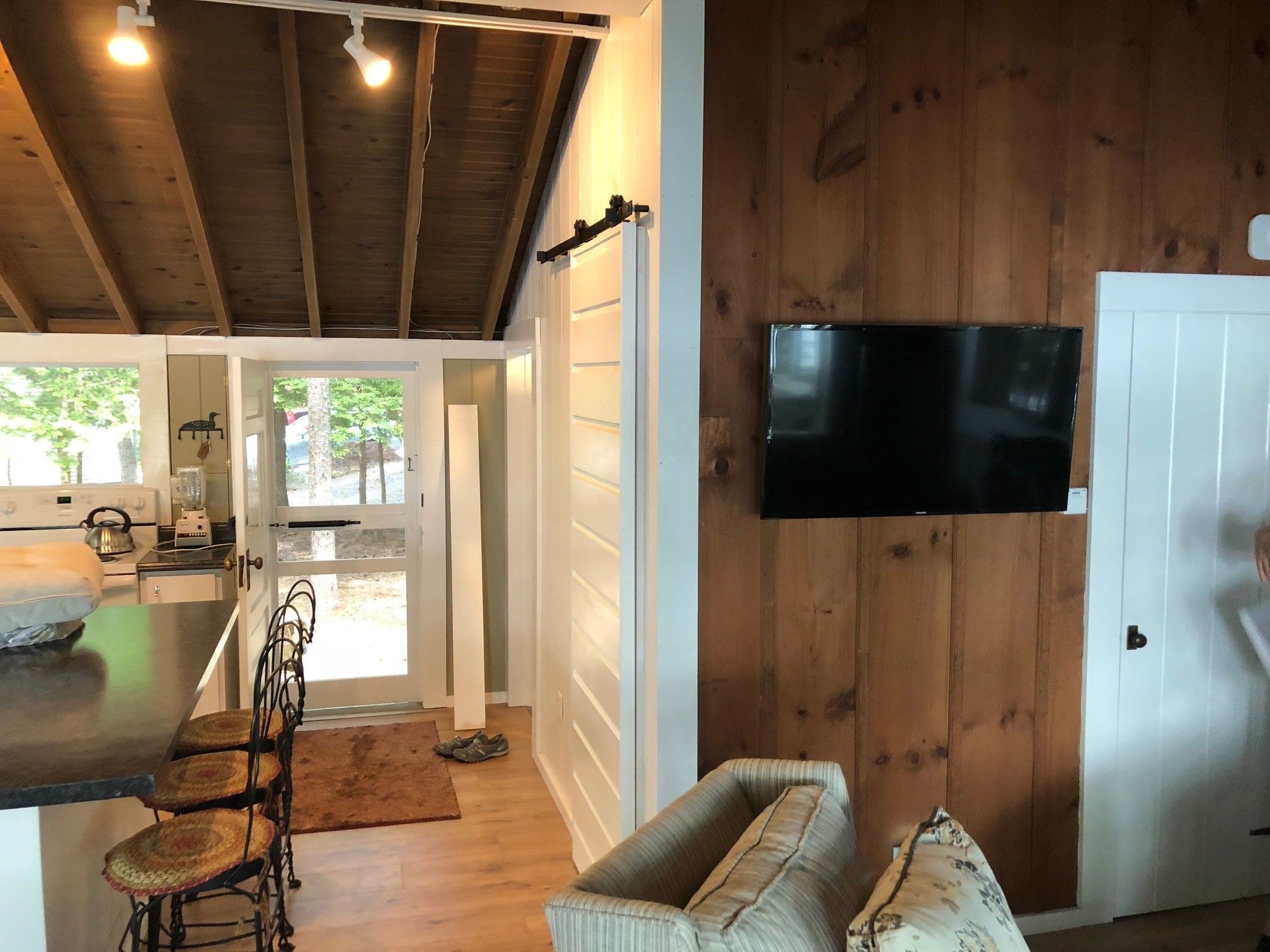 Loon Song Cottage open-concept main living area with knotty pine walls, barn door, wall-mounted TV, kitchen island with barstools, and back door — Meredith, NH