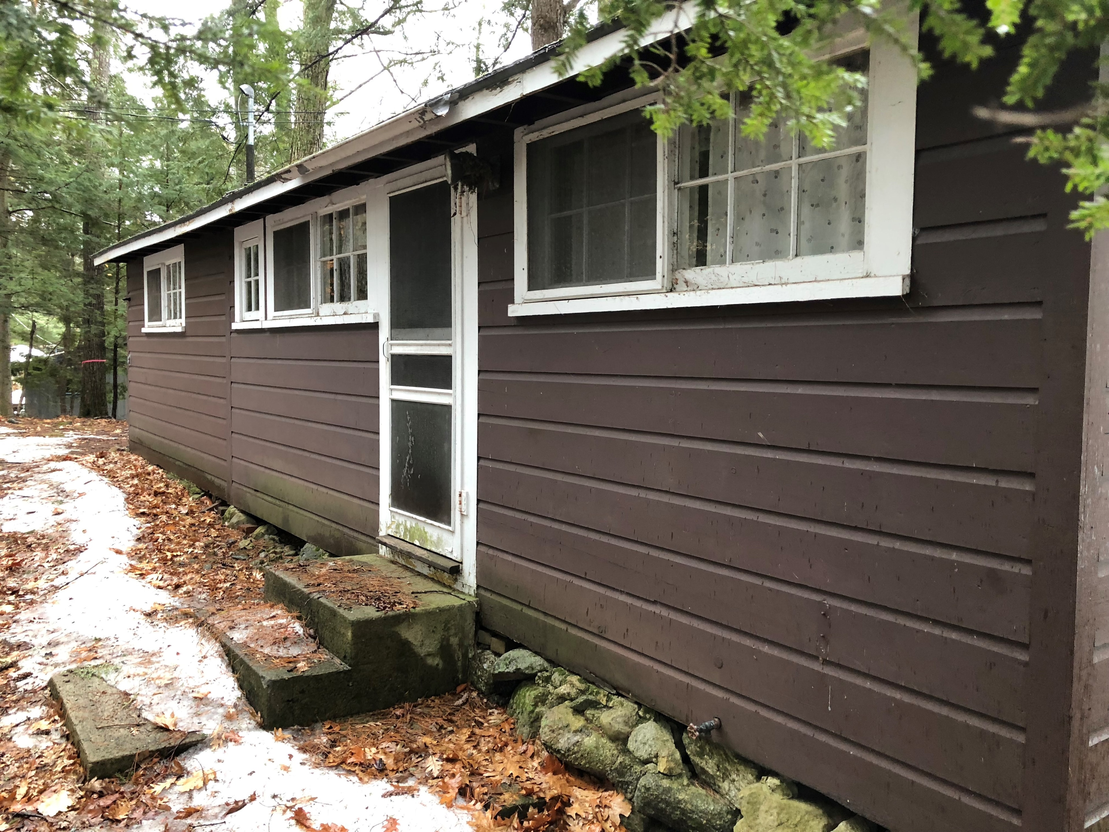 Loon Song Cottage before photo — original dark brown exterior with aging siding, concrete block steps, and overgrown landscaping — Meredith, NH