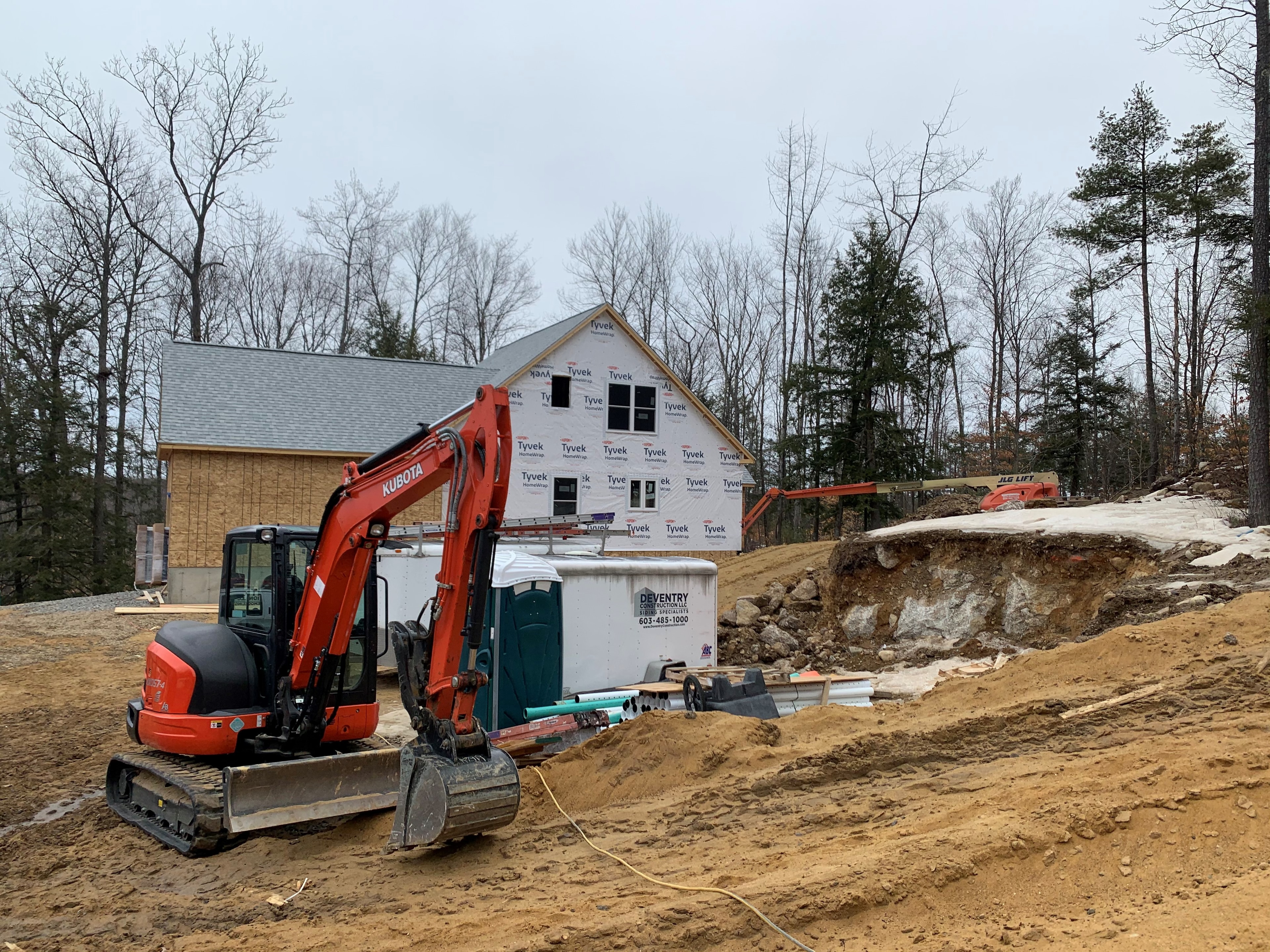 Mason New Build construction progress with Kubota excavator, Tyvek-wrapped framing, Deventry Construction site trailer, and JLG lift — Governor's Crossing, NH